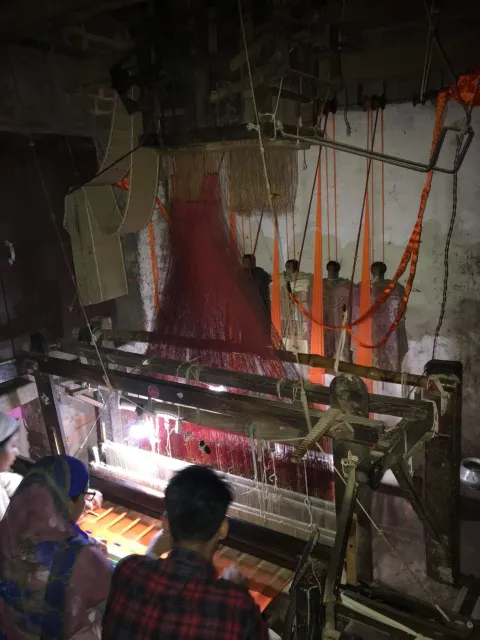 Weavers at a jacquard loom in a dimly lit weaving workshop